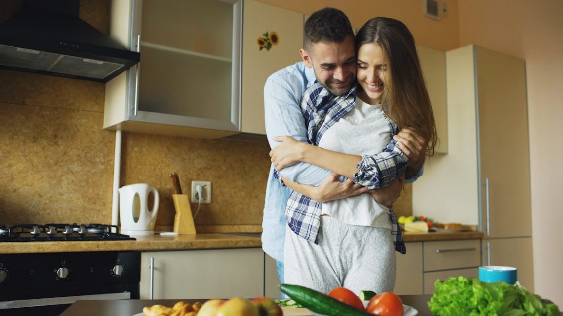 Happy young couple embracing and talking in the kitchen while cooking breakfast at home in the morning